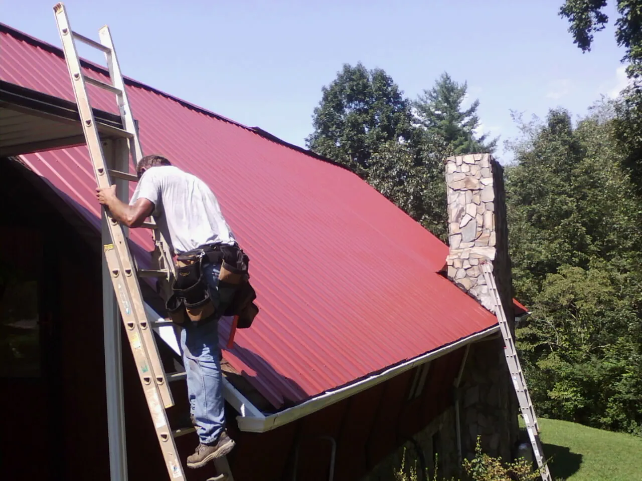 Roofer on a ladder beside a red metal roof and stone chimney — Soffit & Fascia Repair by Roofing Brothers in Lumber Bridge
